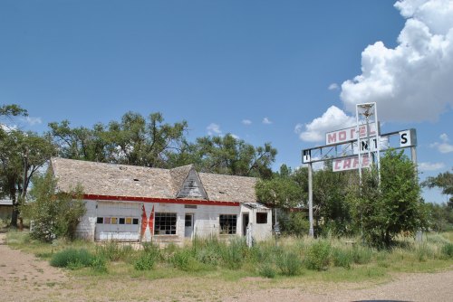 Abandoned motel with a weathered sign, surrounded by overgrown grass and trees under a blue sky with clouds.