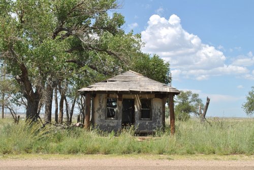 Abandoned, weathered house surrounded by tall grass and trees under a blue sky with fluffy clouds.