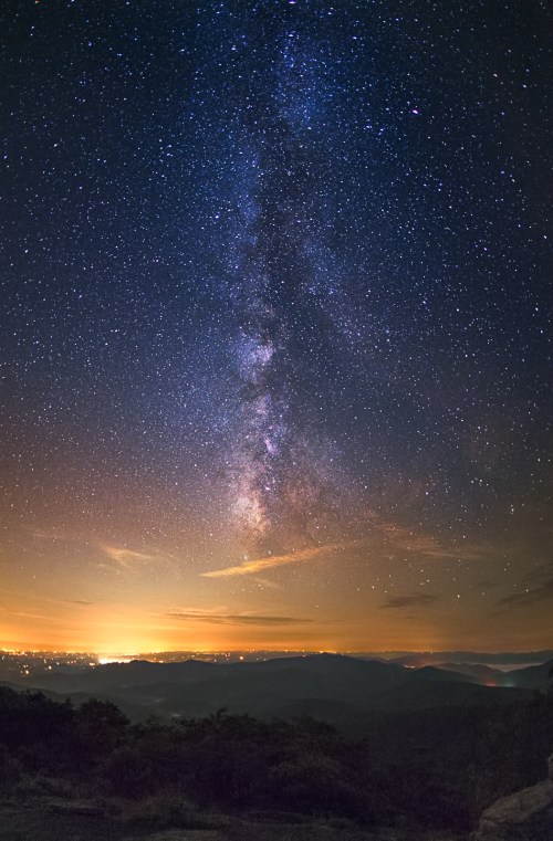 A stunning night sky filled with stars and the Milky Way, above a landscape illuminated by distant city lights.