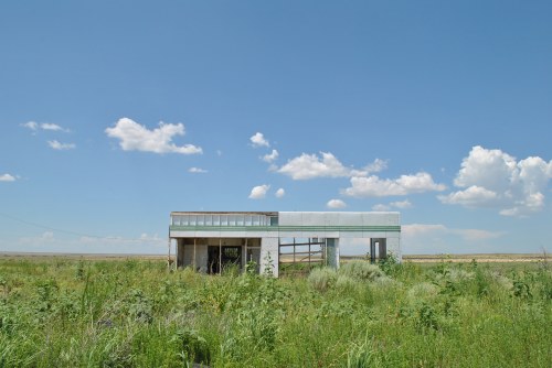 A modern, abandoned building stands in a grassy field under a blue sky with scattered clouds.
