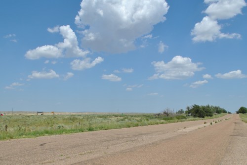 A wide, empty road stretches into the distance under a blue sky with fluffy clouds, surrounded by grassy fields.