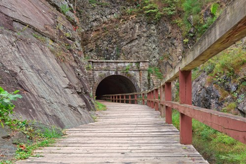 A wooden walkway leads to a dark tunnel, flanked by rocky cliffs and greenery.