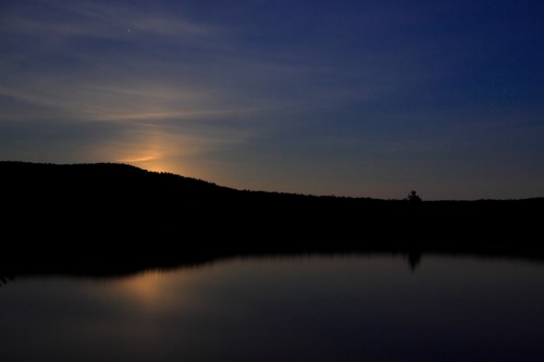 A serene night scene with a calm lake reflecting a faint moonrise behind dark silhouettes of hills.