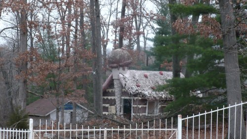 A whimsical, mushroom-shaped house nestled among trees, with a rustic fence in the foreground and autumn leaves scattered.