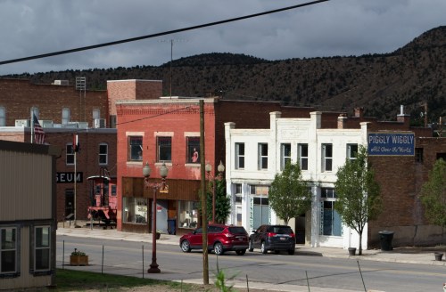 A small town street featuring brick buildings, a museum, and a Piggly Wiggly store, with mountains in the background.