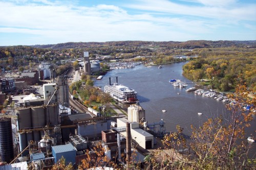 Aerial view of a river with boats, surrounded by trees and buildings, showcasing a scenic landscape in autumn.