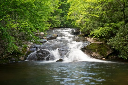A serene waterfall cascades over rocks, surrounded by lush green trees and a calm river.