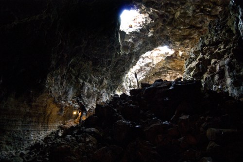 A person explores a dark cave with light streaming in from openings above, illuminating rocky terrain.