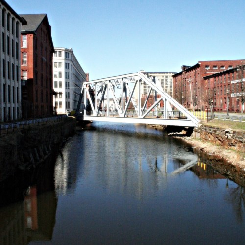 A modern white bridge spans a calm river, flanked by historic brick buildings on either side.