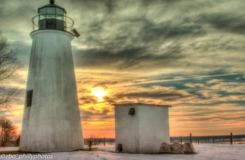 A lighthouse stands beside a small building at sunset, with dramatic clouds and a serene landscape in the background.