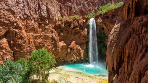 A stunning waterfall cascades into a turquoise pool, surrounded by red rock formations and lush greenery.