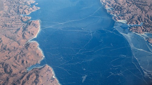 Aerial view of a vast blue lake with visible ice patterns, surrounded by rugged, brown terrain.