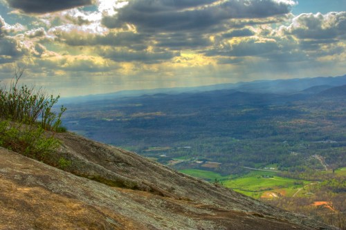 A panoramic view of rolling hills and valleys under a cloudy sky, with sunlight breaking through the clouds.