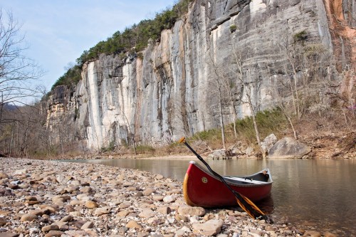 A red canoe rests on a rocky riverbank, with towering cliffs and trees in the background under a clear blue sky.