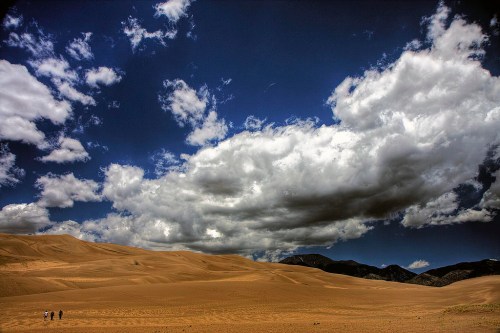Two people walk through a vast desert landscape under a dramatic sky filled with clouds.