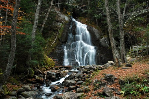 A serene waterfall cascades down rocky cliffs, surrounded by lush trees and autumn foliage.