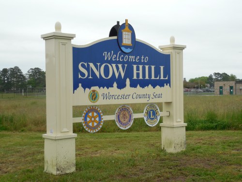Welcome sign for Snow Hill, Worcester County seat, featuring local organization emblems and a scenic background.