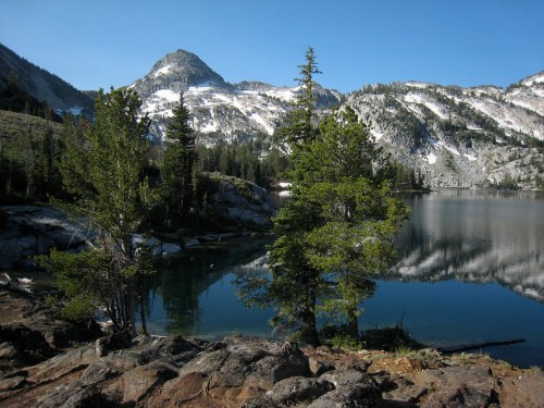 Scenic view of a mountain lake surrounded by trees and rocky terrain, with snow-capped peaks in the background.