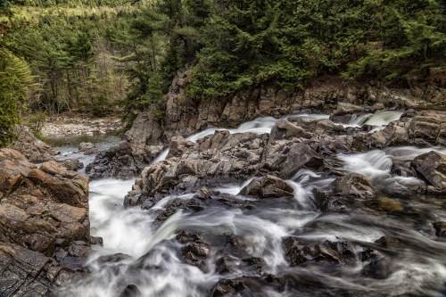 A serene river flows over rocky terrain, surrounded by lush green trees and a peaceful natural landscape.
