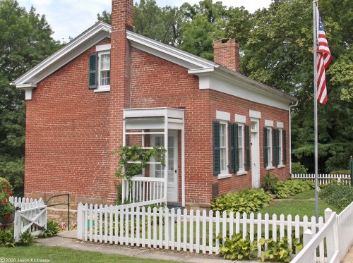 A brick house with white trim, surrounded by a picket fence and greenery, featuring an American flag in the front yard.