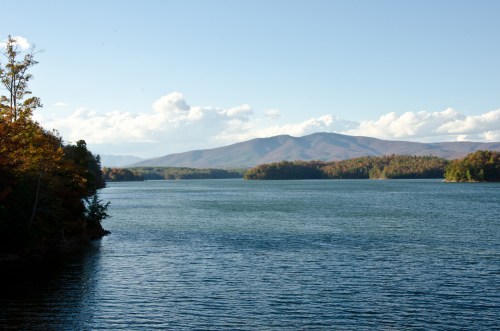 A serene lake surrounded by mountains and trees, with clear blue skies and soft clouds in the background.