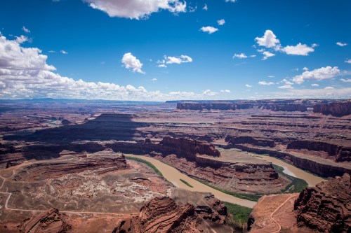 A vast canyon landscape with a winding river, under a bright blue sky dotted with clouds.