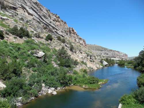 A serene river flows through a rocky landscape, surrounded by lush greenery and clear blue skies.