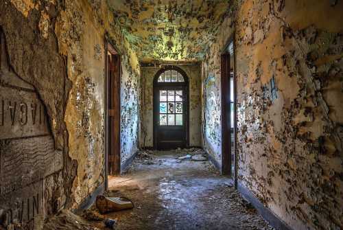 A dimly lit, abandoned hallway with peeling walls and a door at the end, covered in dust and debris.