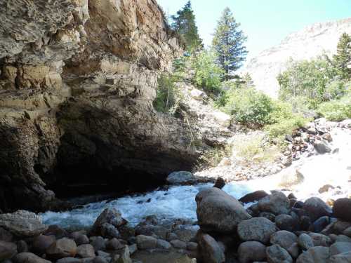 A rocky riverbank with rushing water, surrounded by lush greenery and a cave entrance in a mountainous area.