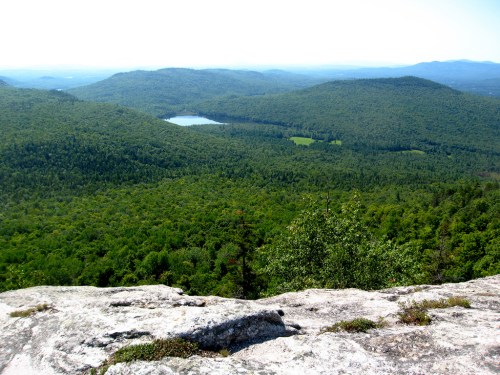 A panoramic view of lush green mountains and a small lake, seen from a rocky overlook on a clear day.