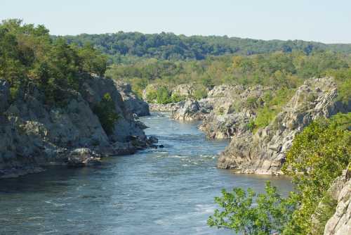 A serene river flows between rocky cliffs, surrounded by lush greenery under a clear blue sky.