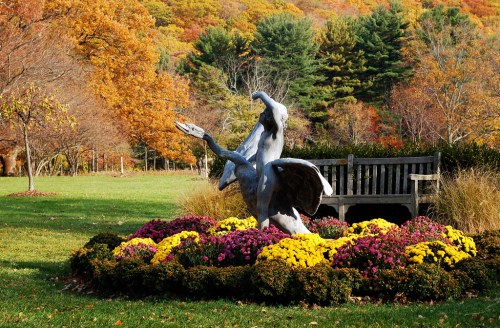 A silver statue of a bird in flight surrounded by colorful flowers and autumn foliage, with a bench nearby.