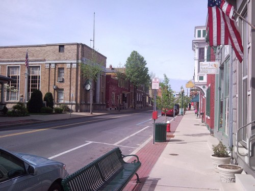 A quiet street scene featuring buildings, trees, and American flags, with a bench along the sidewalk.