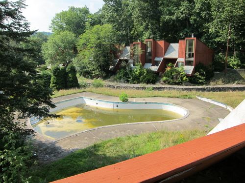 Abandoned swimming pool filled with green water, surrounded by overgrown grass and modernist-style buildings in the background.