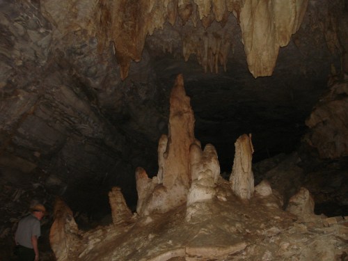 A cave interior featuring stalagmites and stalactites, with a person standing in the background for scale.