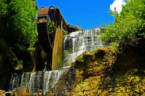 A waterwheel beside a cascading waterfall, surrounded by lush greenery and a clear blue sky.