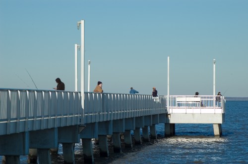 A pier extends over calm water, with people fishing and relaxing under a clear blue sky.