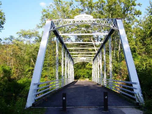 A white metal bridge spans a path surrounded by lush green trees under a clear blue sky.