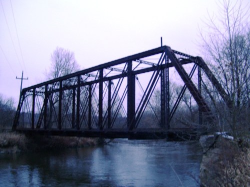A rusty, old railway bridge spans a calm river, surrounded by bare trees and a cloudy sky.