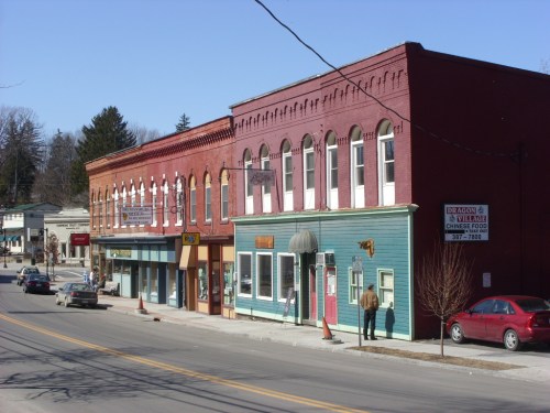 A small town street featuring colorful storefronts and a clear blue sky, with a few people walking along the sidewalk.