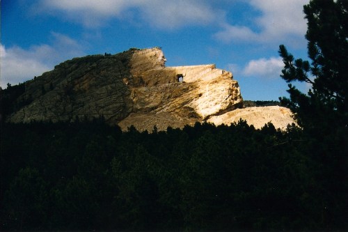 A rugged mountain peak illuminated by sunlight, surrounded by dense green trees and a blue sky with clouds.