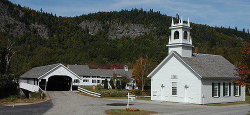 A white church with a bell tower next to a covered bridge, surrounded by trees and mountains in a rural setting.