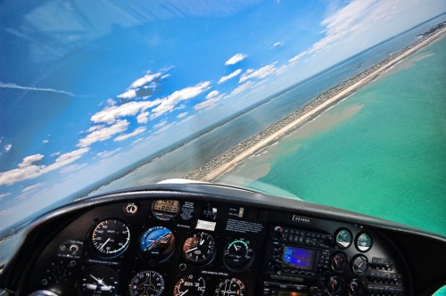 Cockpit view of a small aircraft flying over a coastal area with turquoise waters and sandy shores under a blue sky.