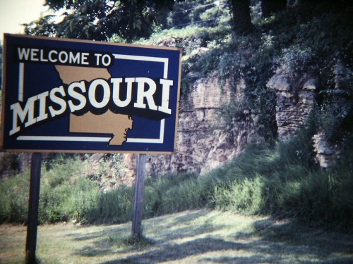 A weathered sign reading "Welcome to Missouri" stands in front of a rocky hillside and grassy area.