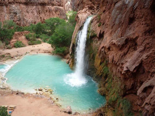 A stunning waterfall cascades into a turquoise pool surrounded by red rock cliffs and lush greenery.