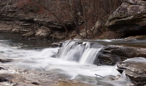 A serene waterfall cascades over rocky ledges, surrounded by trees and a calm river.