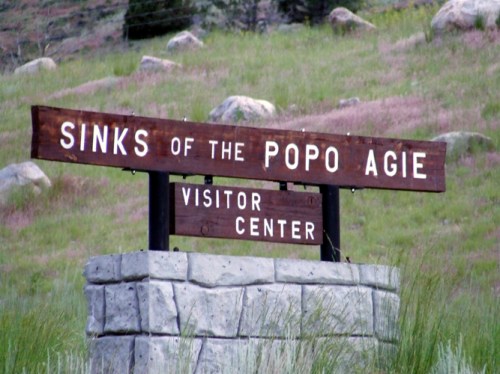 Wooden sign for "Sinks of the Popo Agie Visitor Center" in a grassy area with rocks in the background.