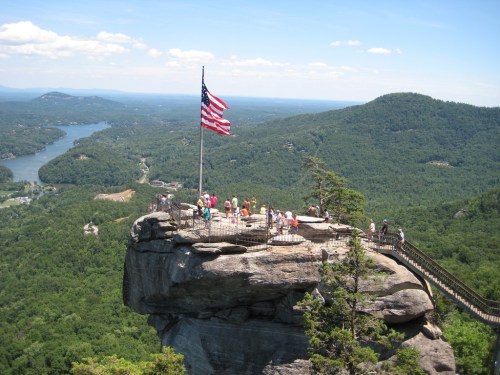A scenic view from a rocky overlook with an American flag, surrounded by lush green mountains and a winding river below.