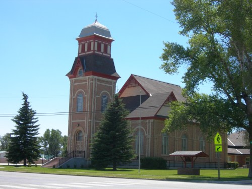 A brick church with a tall tower, surrounded by trees and a clear blue sky, located near a road with a pedestrian crossing sign.