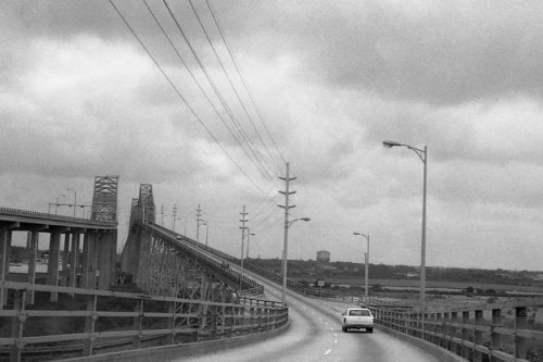 A black and white photo of a winding road leading to a bridge under a cloudy sky, with power lines along the side.